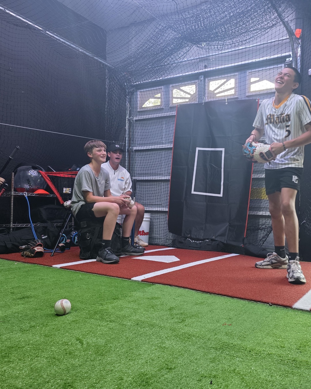 Three athletes laughing inside the Garage Lab batting cage in Stafford. One stands with a glove; two sit on a bench.